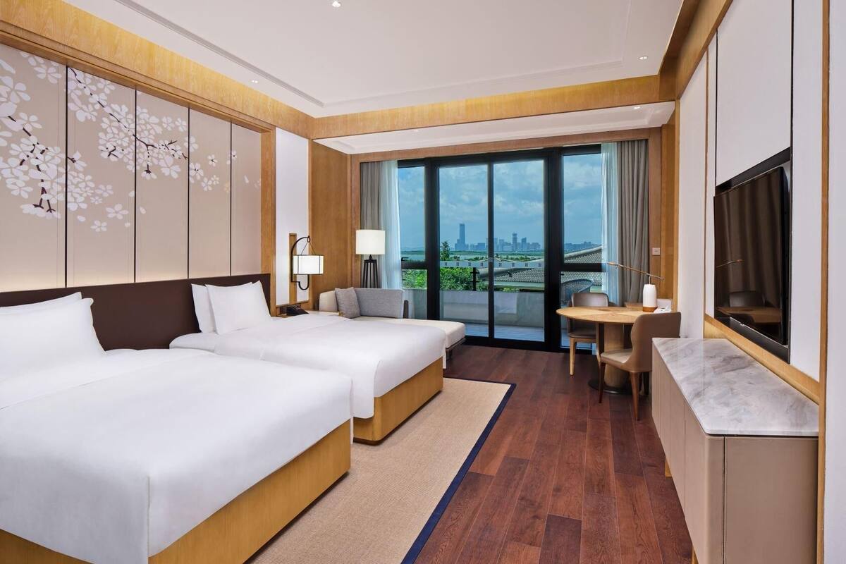A hotel room with a large bed, a desk, a chair, a TV, and a view of the city.