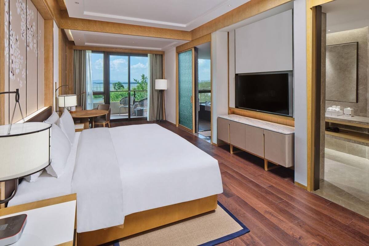 A hotel room with a bed, a TV, a balcony with a view, and a desk with a lamp.