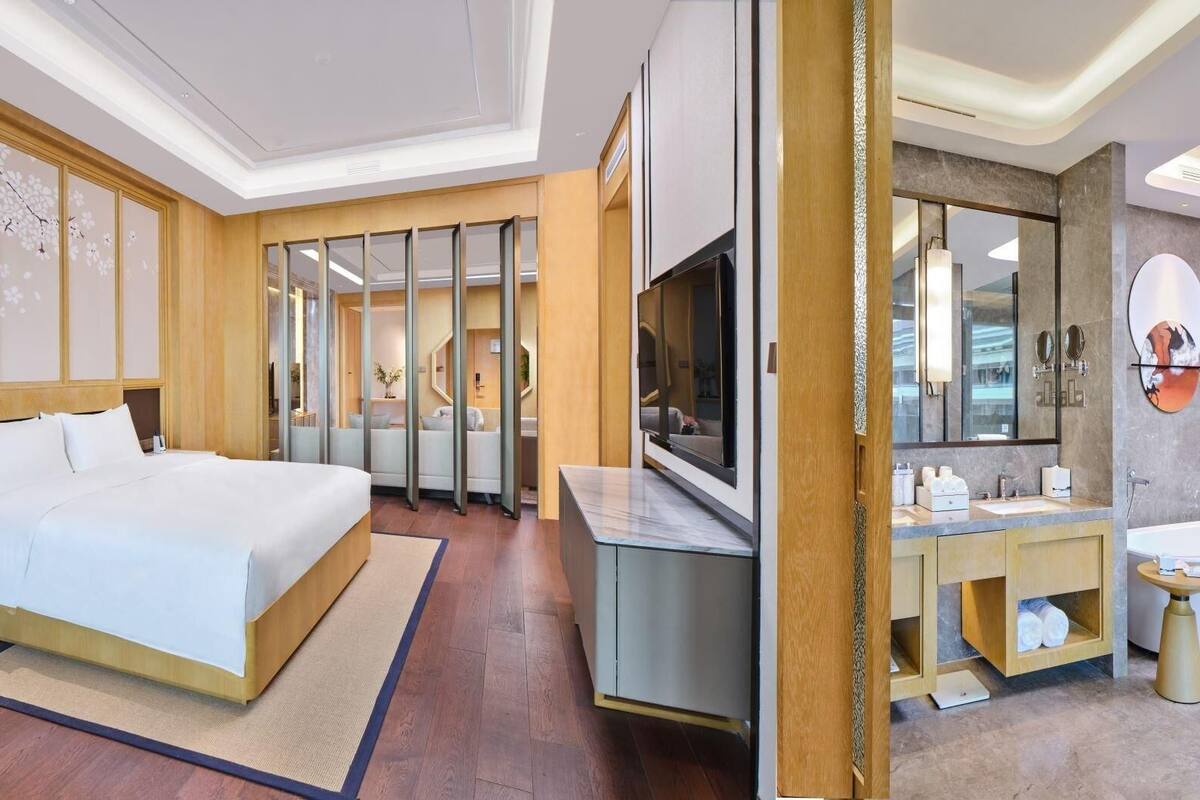 A modern hotel room with a large bed, a TV, a bathroom with a mirror, and a wooden floor.