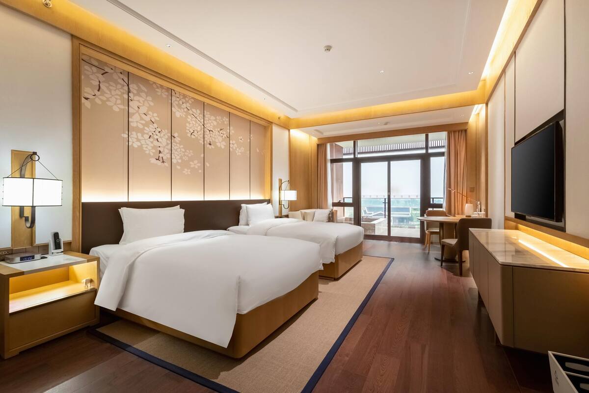 A hotel room with two beds, a desk, a TV, and a large window with a view.