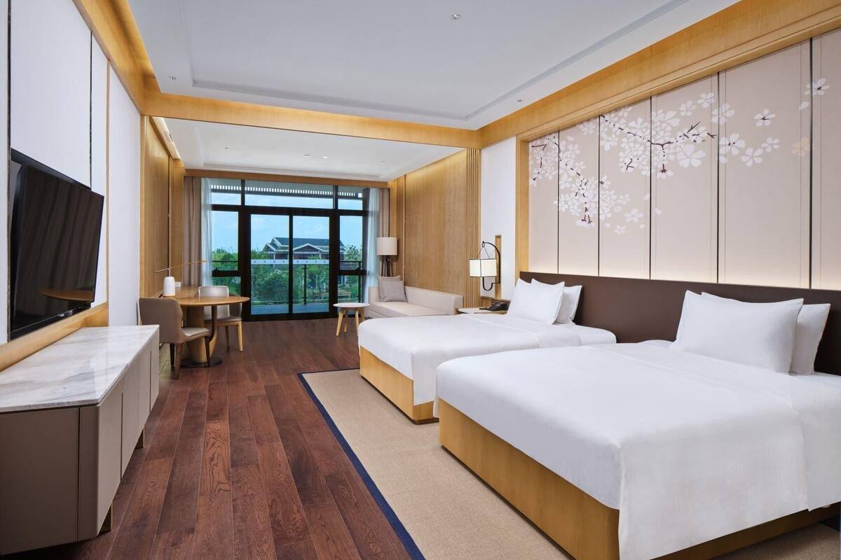 A hotel room with two beds, a desk, a TV, and a large window.