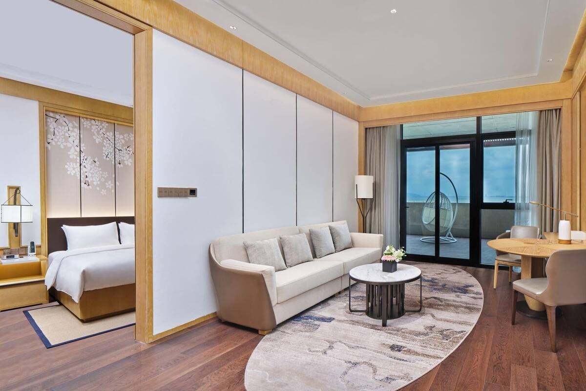 A hotel room with a bed, a sofa, a dining table with chairs, a small coffee table, and a large window with a view.
