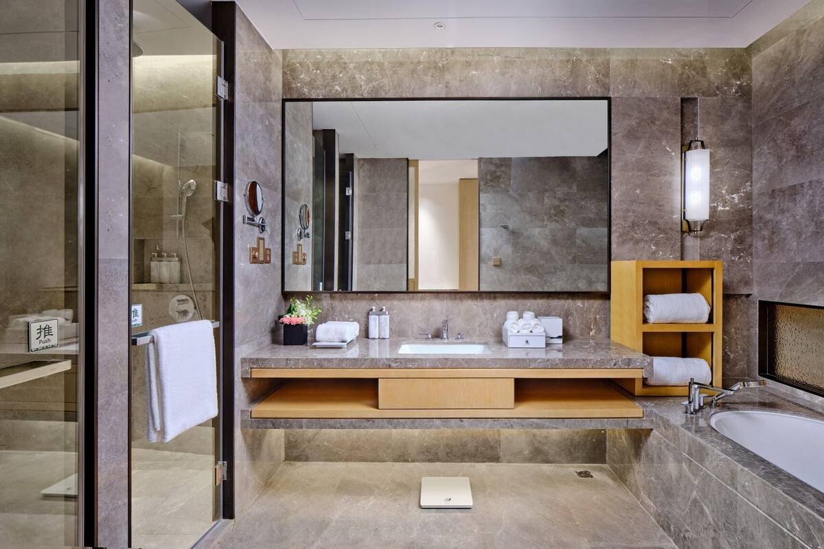 A modern bathroom with a large mirror, a sink, a shower, and a wooden shelf with towels.