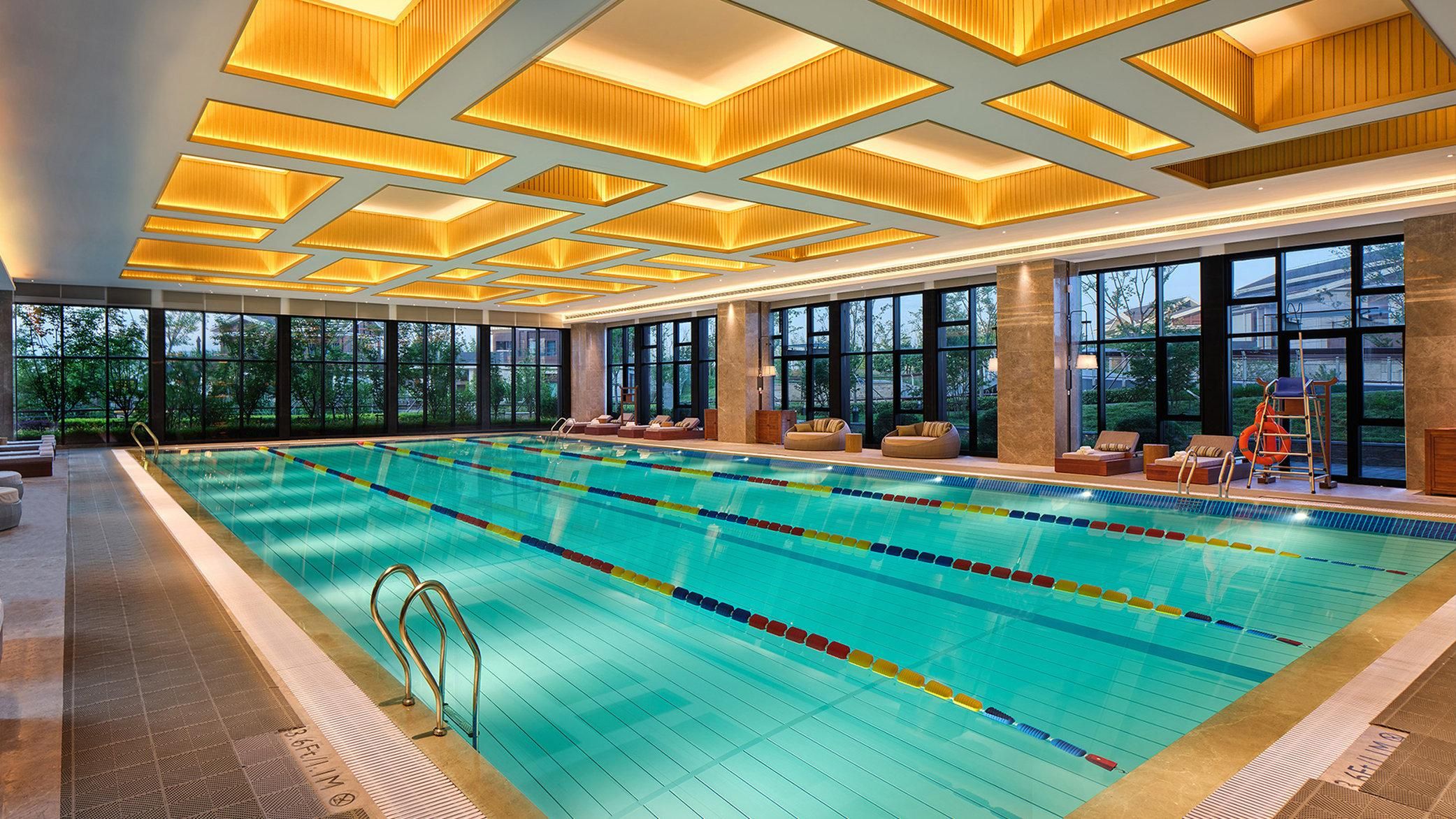 Une grande piscine intérieure avec des couloirs clairement délimités, un espace salon aménagé en contrebas avec des coussins, et de grandes fenêtres offrant une vue sur l’extérieur.