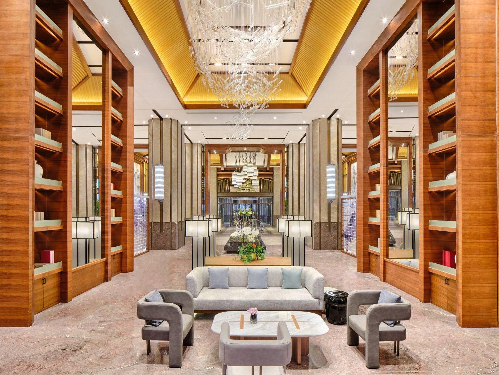 A modern hotel lobby with a central seating area, wooden bookshelves, and a chandelier.