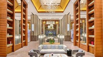 A modern hotel lobby with a central seating area, wooden bookshelves, and a chandelier.