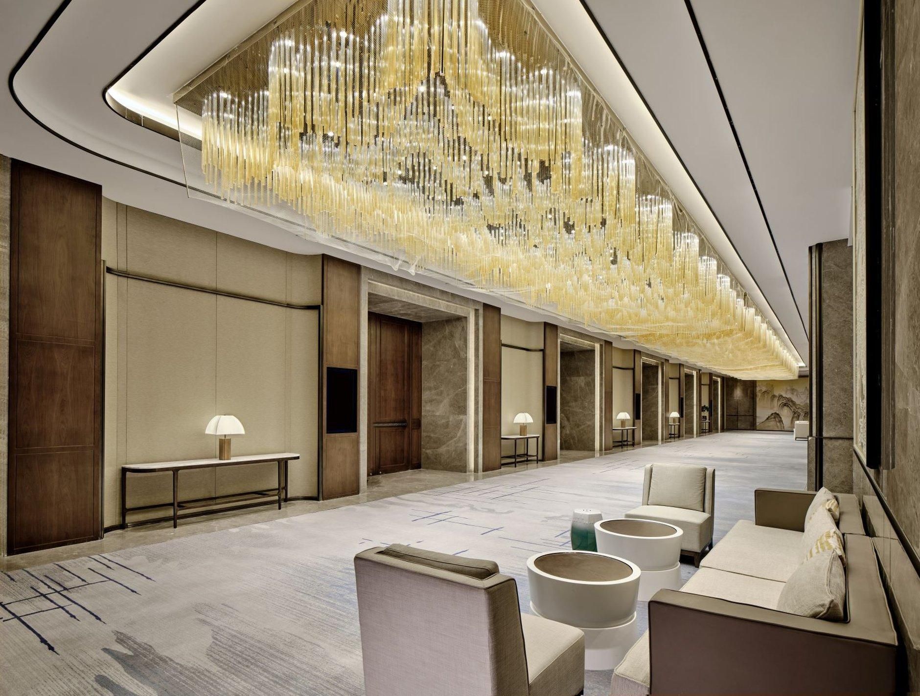 A hotel lobby with a long corridor, seating areas, and a decorative ceiling light fixture.