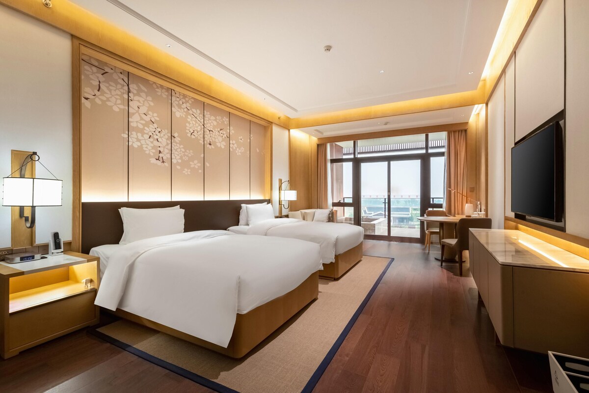 A hotel room with two beds, a desk, a TV, and a large window with a view.
