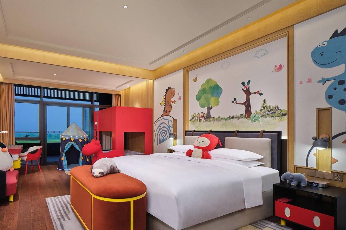 A hotel room with a large bed, a red play tent, a desk with a chair, and a window with a view.