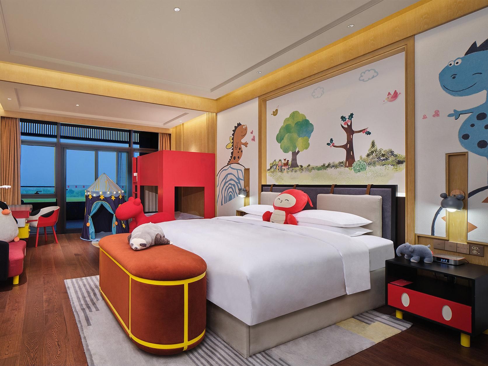 A hotel room with a large bed, a red play tent, a desk with a chair, and a window with a view.