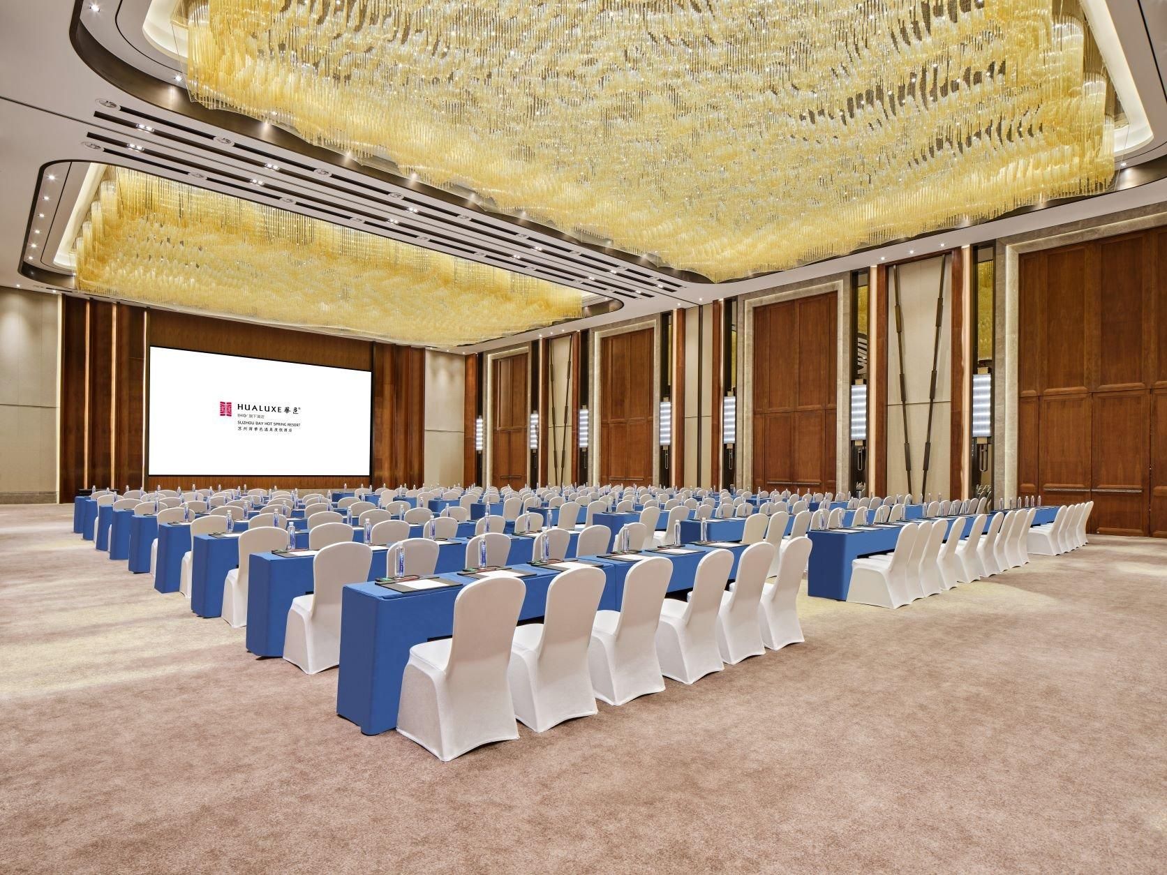 Un salón de conferencias con filas de mesas y sillas, una pantalla grande y un techo decorativo.