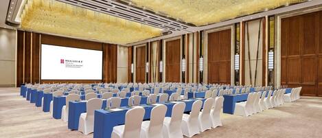 Un salón de conferencias con filas de mesas y sillas, una pantalla grande y un techo decorativo.
