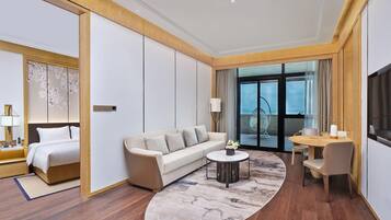 A modern hotel room with a bed, sofa, round coffee table, and a dining area with a chair and table.