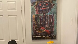 Un coin repas avec une table noire et deux chaises noires, un saladier de fruits sur la table et un tableau de Jean Dubuffet accroché au mur.