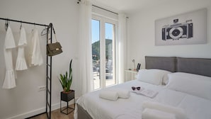 A hotel room with a bed, a plant, a camera on the wall, and a window with a view of the outside. - Apartment Blù (Dubrovnik)