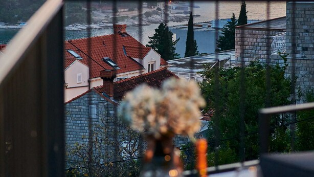 A balcony with a table set for dining, overlooking a coastal town with buildings and a view of the sea. - Apartment Blù (Dubrovnik)
