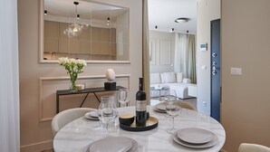 A modern dining area with a round table set for four, a wine bottle, and a vase of flowers. A large mirror and a sofa are visible in the background. - Apartment Blù (Dubrovnik)