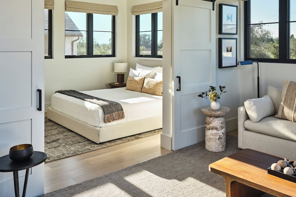 A modern bedroom with a bed, a sofa, and a coffee table. There are windows with views of greenery.