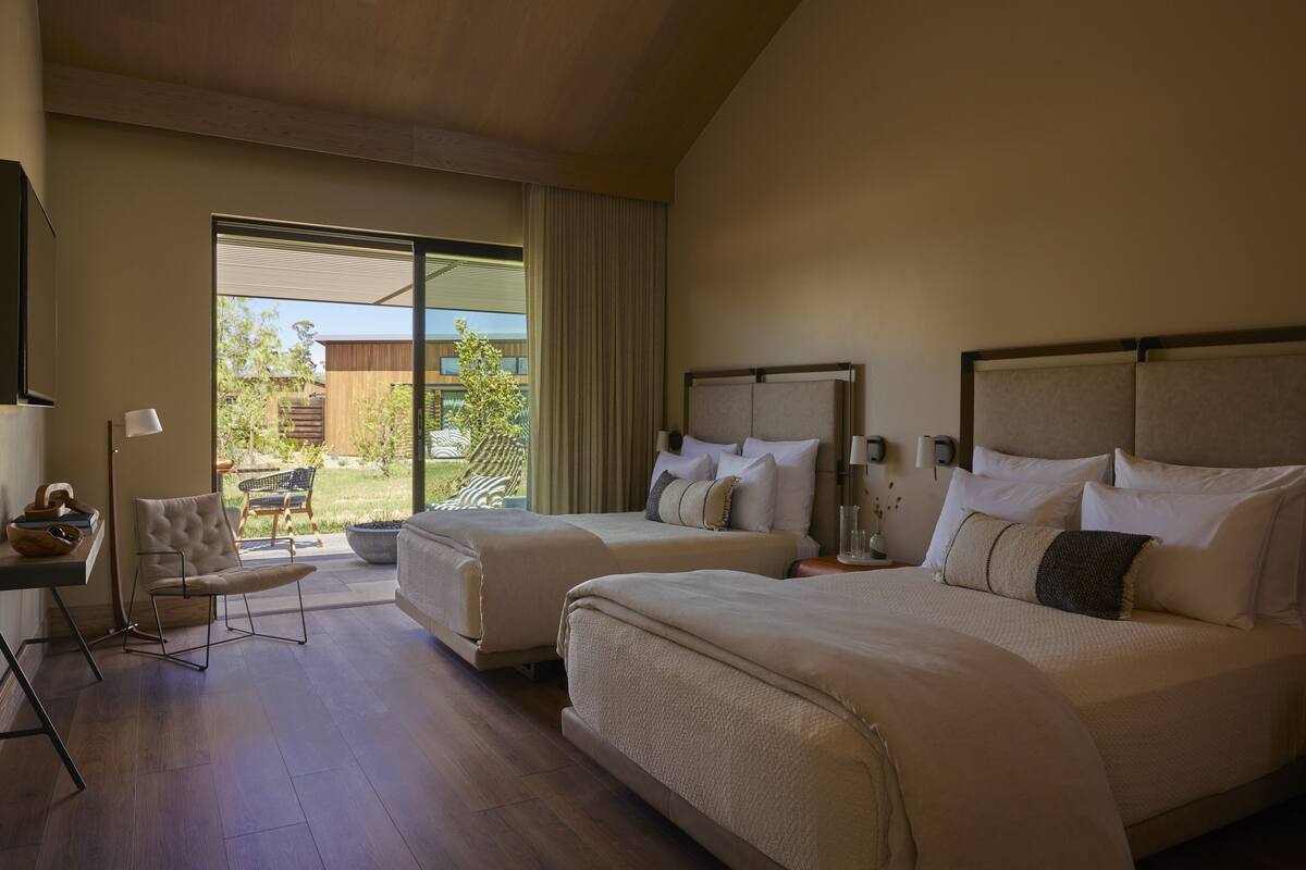 A hotel room with two beds, a desk, a chair, and a large window with a view of the outdoors.