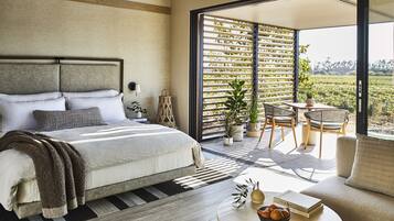 A modern bedroom with a large bed, a small table with books and a bowl of fruit, and a balcony with a view of a vineyard.