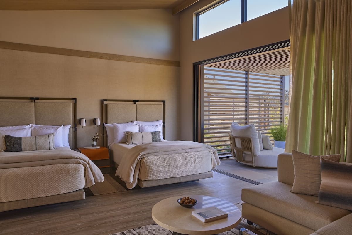 A modern hotel room with two beds, a sofa, a coffee table, and a large window with blinds.