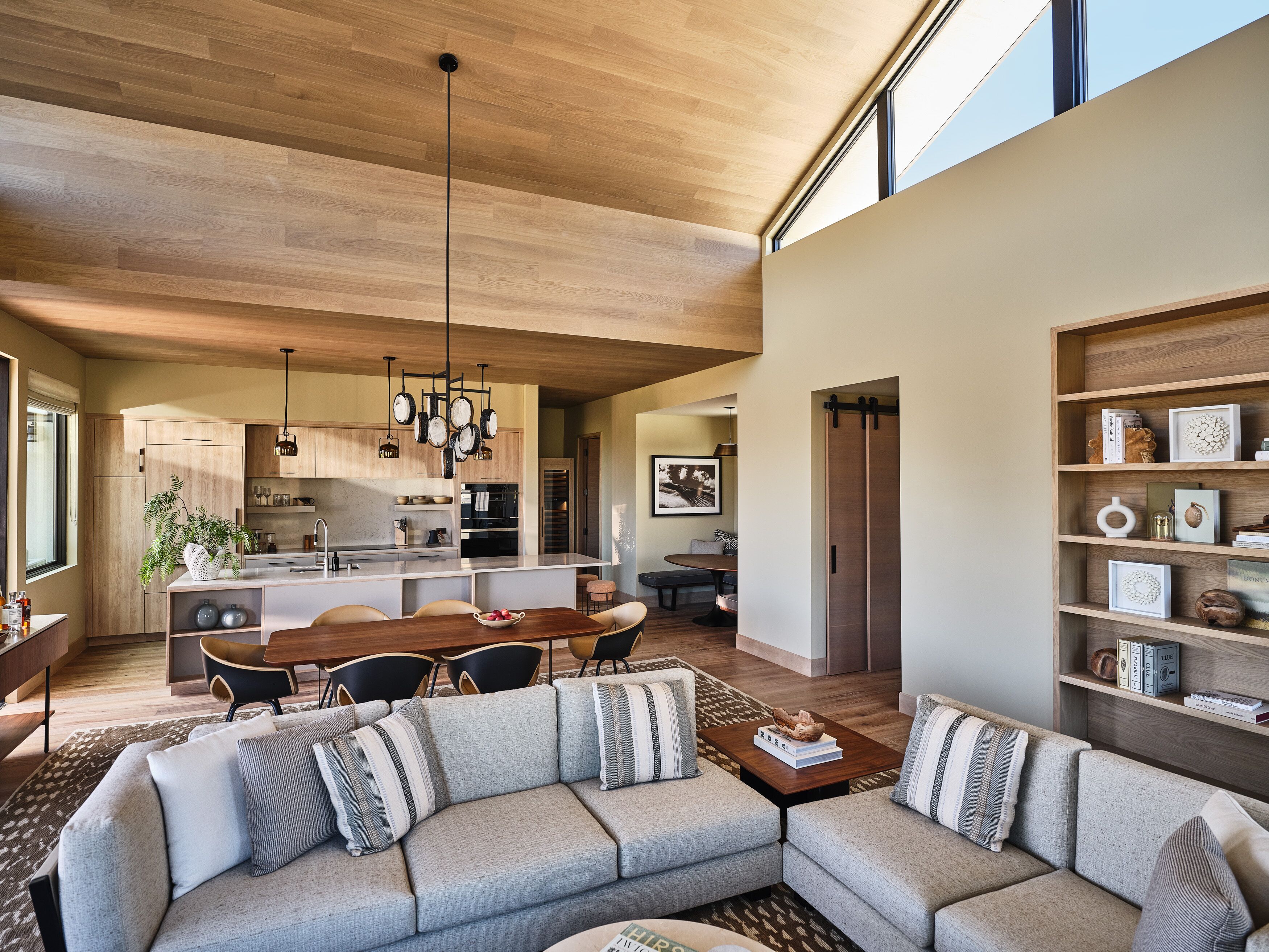 A modern living room with a sectional sofa, a dining area, and a kitchen with wooden cabinetry and pendant lighting.