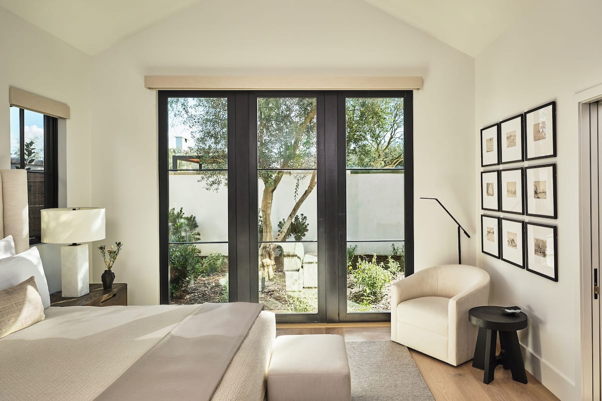 A bedroom with a large window, a bed, a chair, a desk, and framed pictures on the wall.