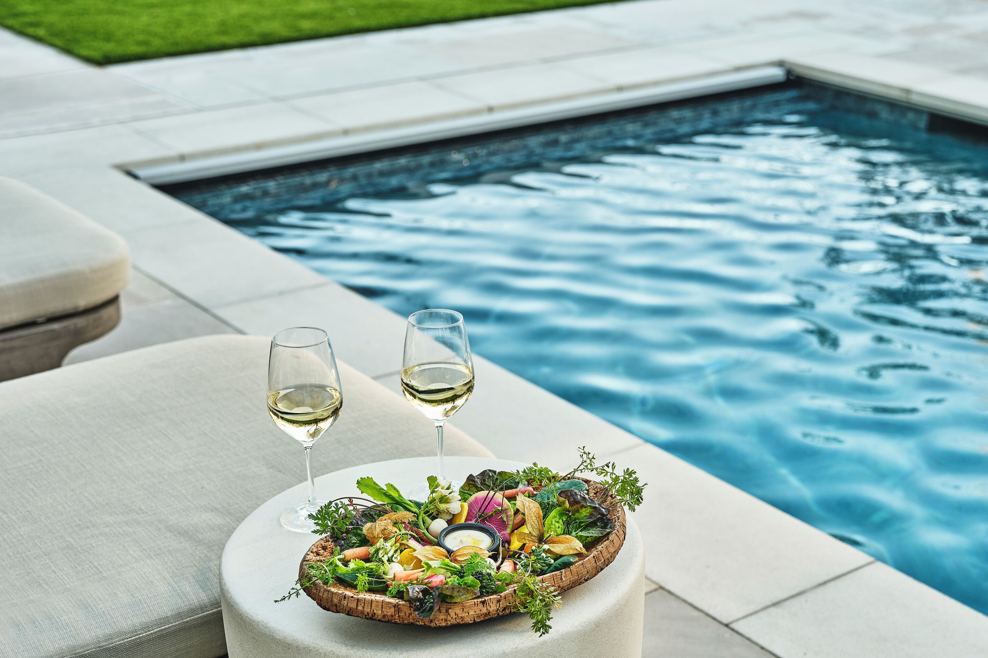 Ein Poolbereich mit einem runden Holztisch, auf dem ein Salat und zwei Gläser Weißwein stehen.