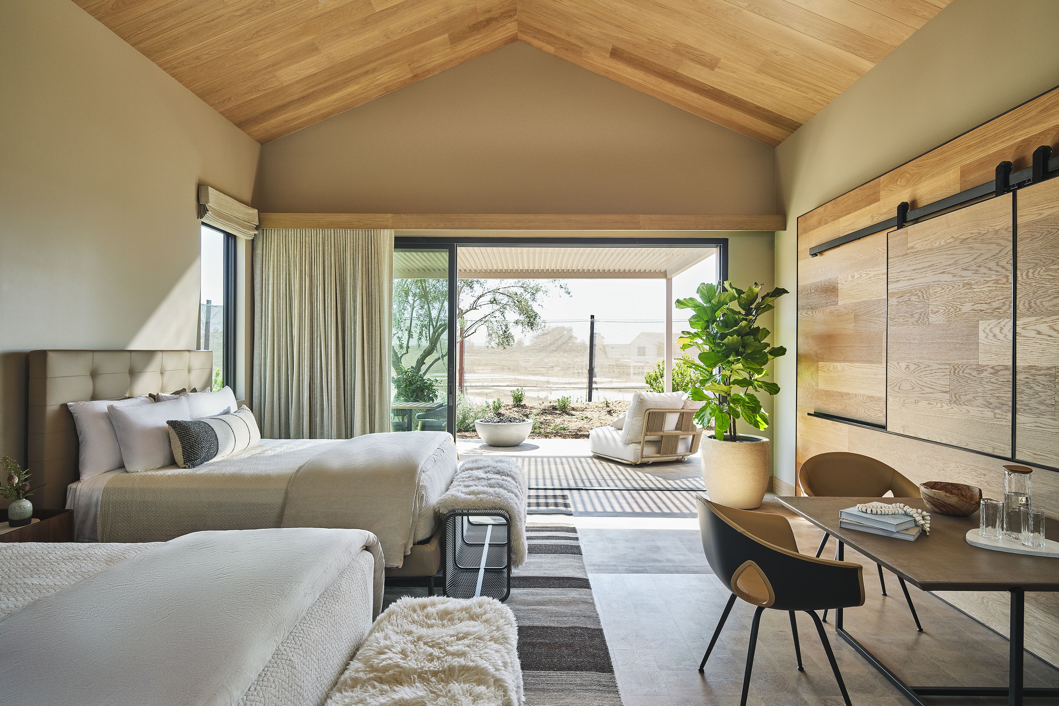 A modern hotel room with two beds, a dining area, and a large sliding door leading to an outdoor patio.