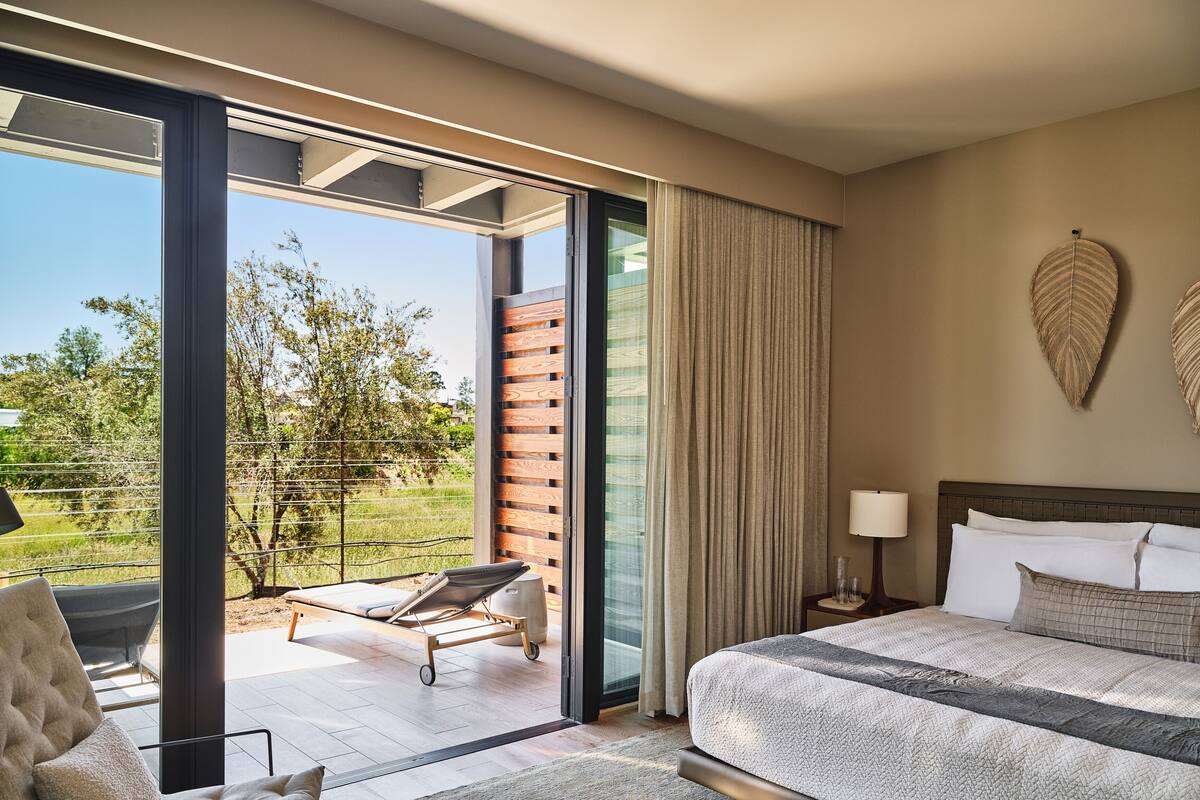 A modern hotel room with a large bed, a chair, a small table with a lamp, and a view of the outdoors.