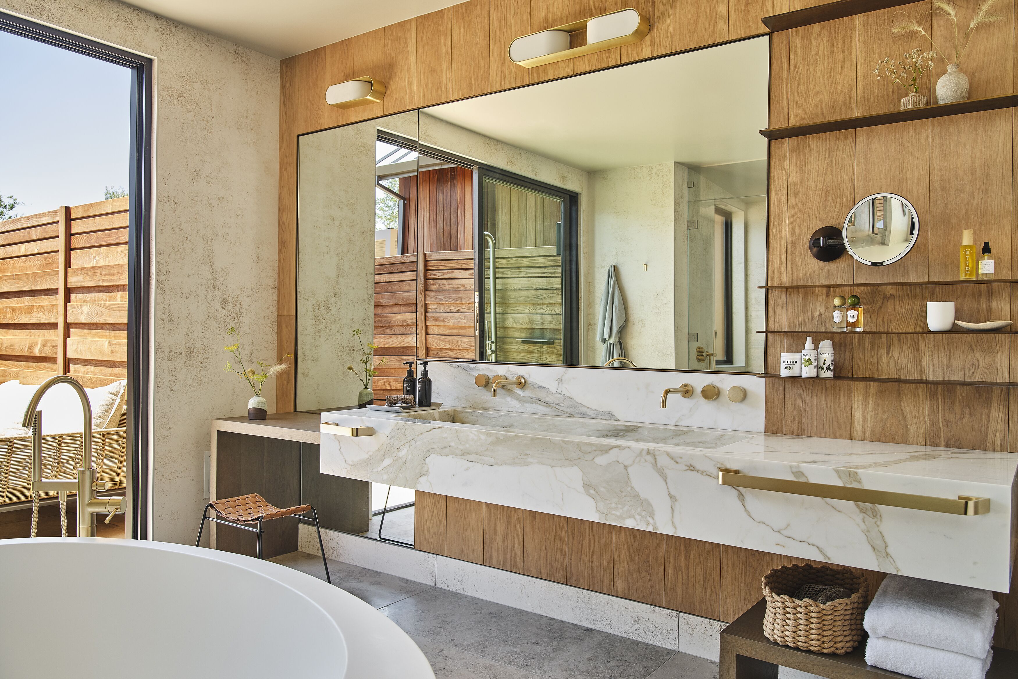 A modern bathroom with a large mirror, marble countertop, wooden accents, and a view of the outdoors.