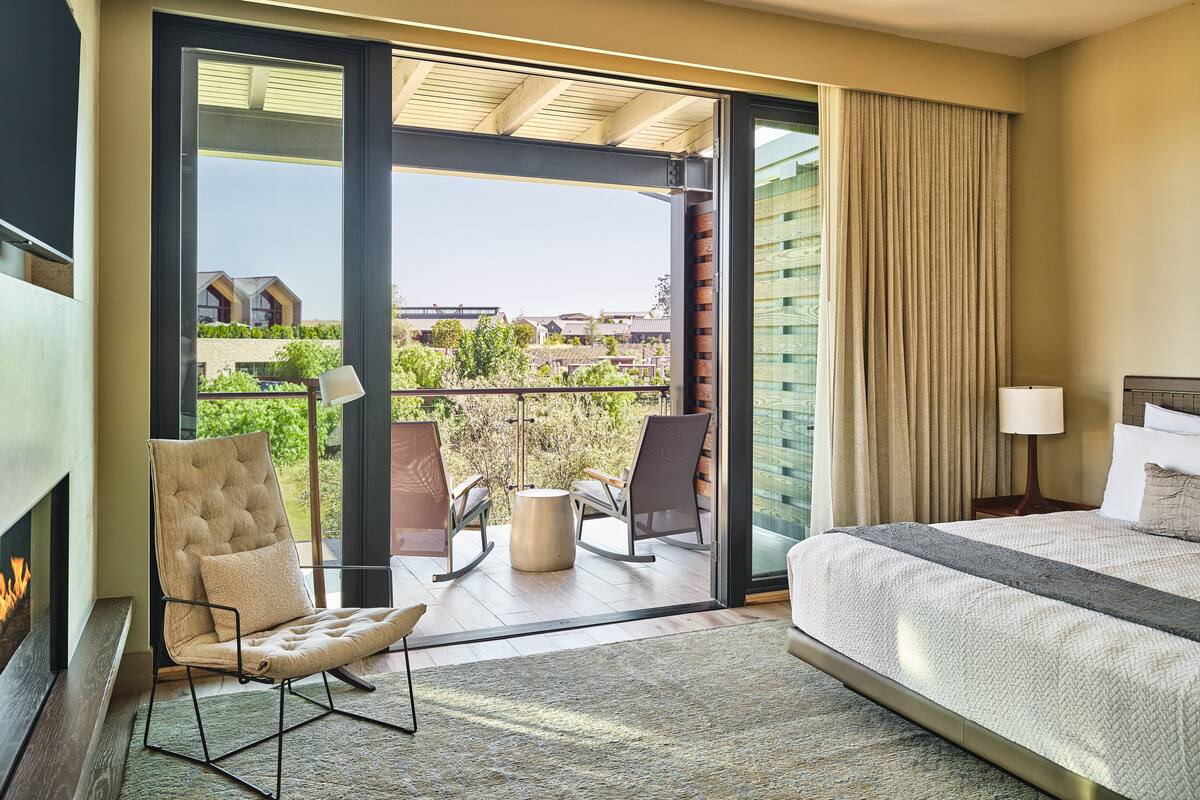 A hotel room with a bed, a chair, a table, and a view of the outdoors.