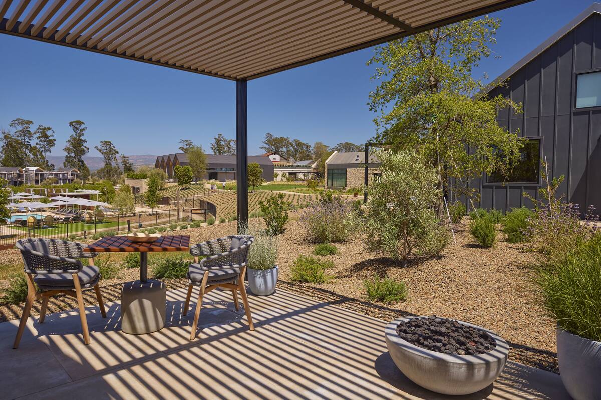 An outdoor patio with a table and chairs, surrounded by a garden with various plants and a view of the surrounding landscape.