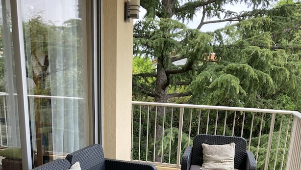 Een balkon met rieten meubilair, waaronder een bank en twee fauteuils, en een glazen deur die uitkomt op een tuin.