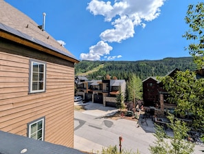 A wooden building with a window, a paved area, and a view of a forested mountain range. - Summer in River Run | Pool, Hot Tubs, Views (Keystone)