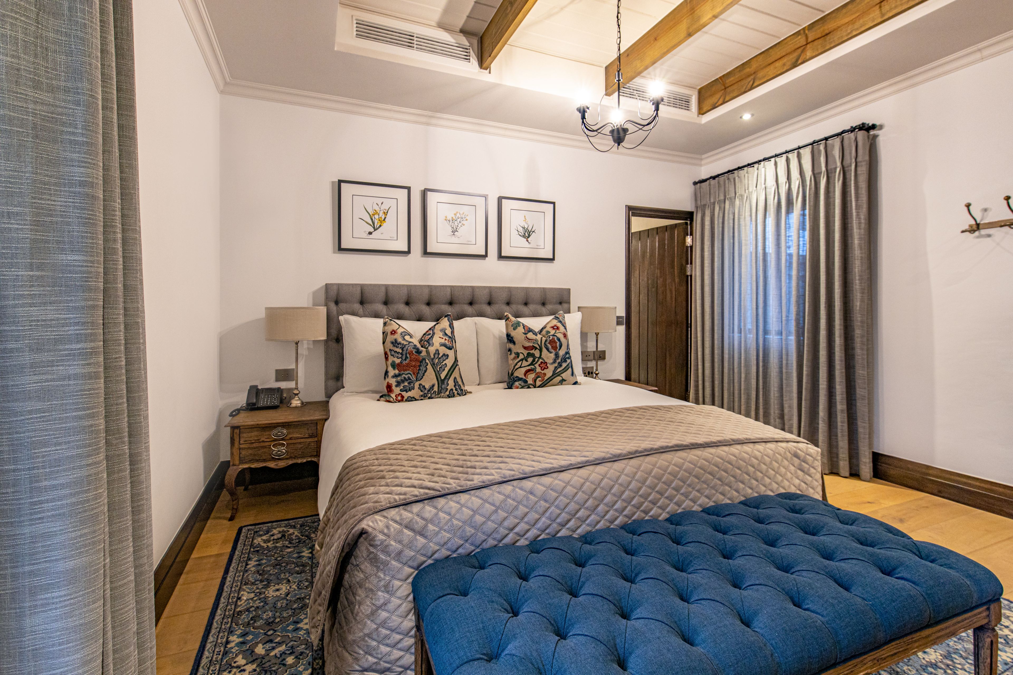 Quarto com cama grande, um banquinho azul estofado, mesa de cabeceira de madeira e mesa lateral com abajur.