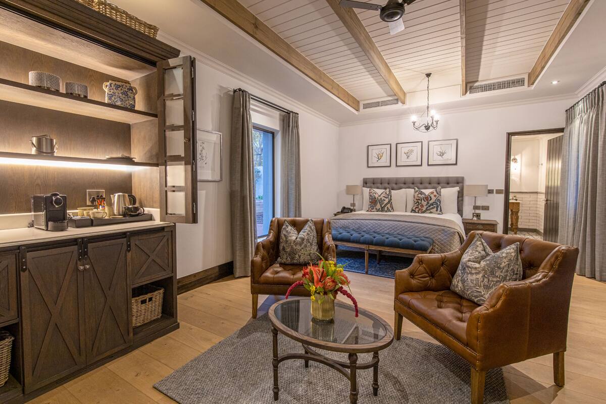 A modern hotel room with a wooden ceiling, a large bed, a sitting area with leather chairs, a glass coffee table, and a kitchen area with a countertop and cabinets.