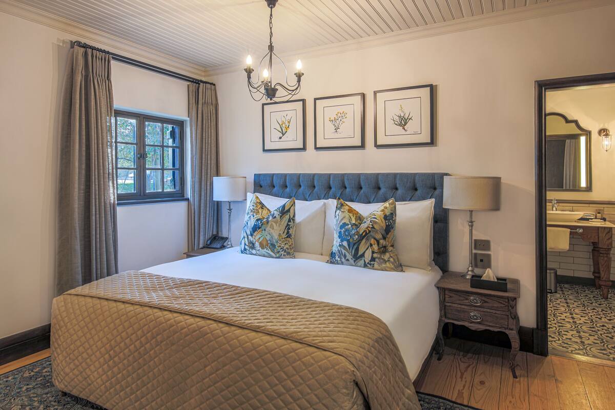 A hotel room with a large bed, a nightstand, a mirror, and framed botanical prints on the wall.
