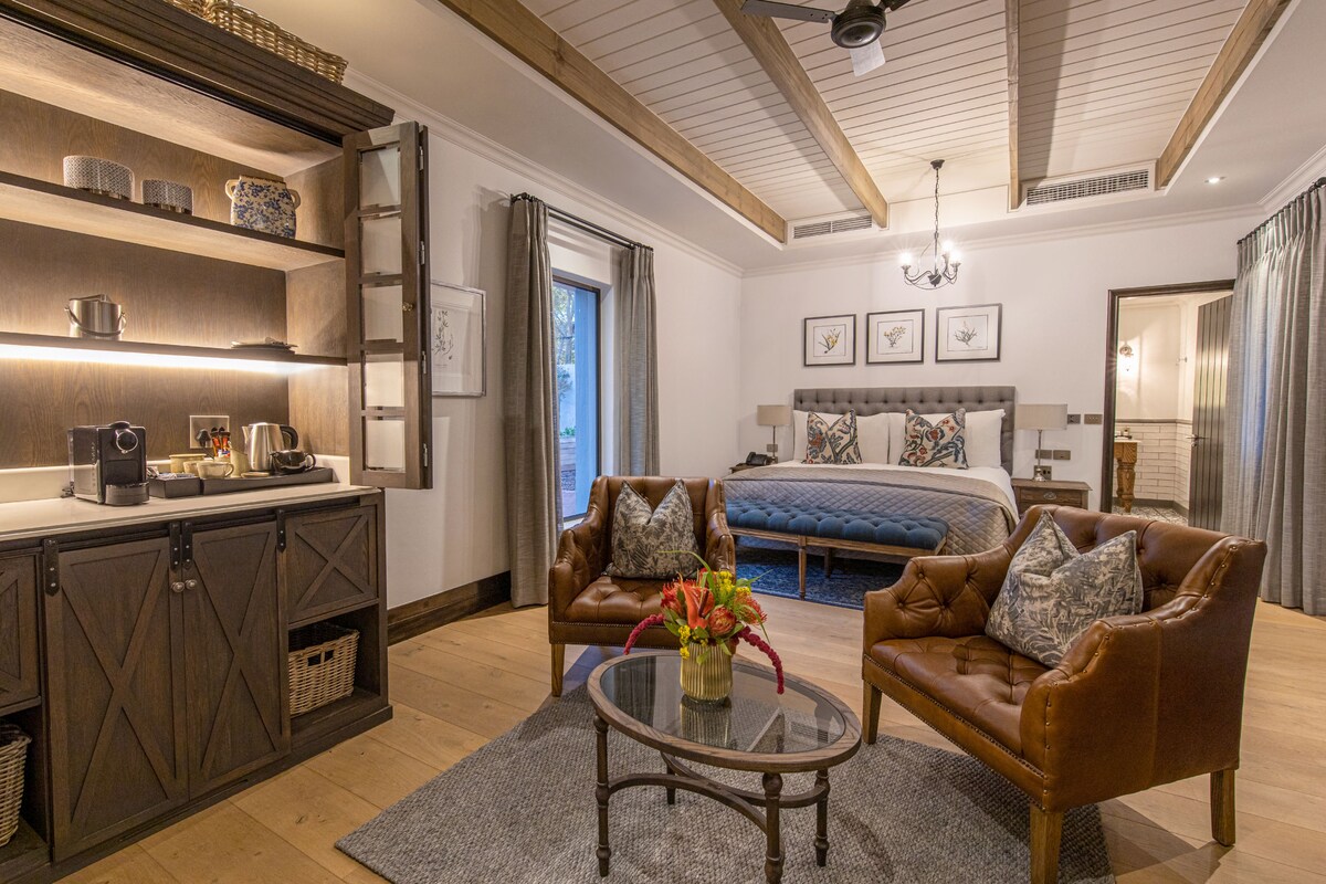 A modern hotel room with a wooden ceiling, a large bed, a sitting area with leather chairs, a glass coffee table, and a kitchen area with a countertop and cabinets.