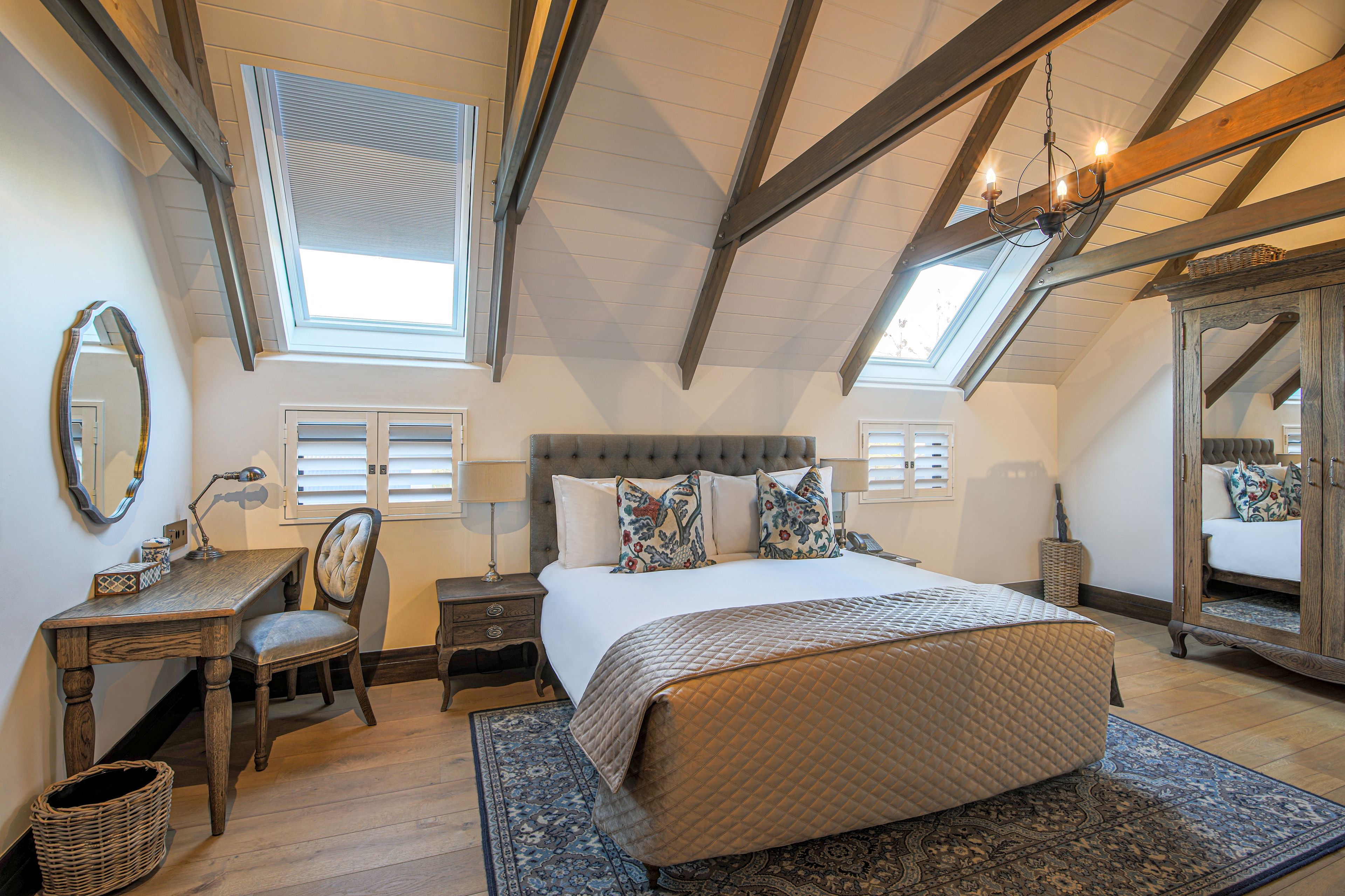 A bedroom with a large bed, a desk, and a chair. There are two windows with blinds, a chandelier, and a mirror on the wall.