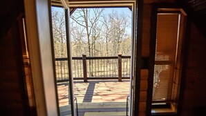 Una terraza de madera con baranda metálica, que conduce a una puerta con vistas a árboles y un cielo despejado.