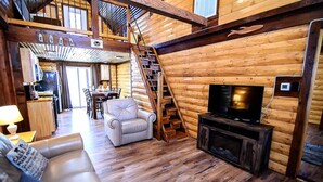 Interior de una cabaña de troncos con escalera de madera, un sofá de cuero, un televisor y un comedor.