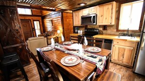 Interior de una cabaña de madera con una mesa de comedor preparada para cuatro personas, armarios de madera, un microondas y un refrigerador.