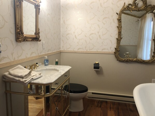 Un baño con un espejo en marco dorado, un lavamanos blanco con encimera de mármol, un inodoro y una silla blanca.