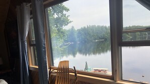 A room with a wooden chair and a bed, a window overlooking a lake with trees and a dock.