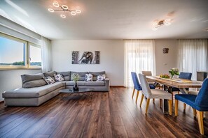 A modern living room with a grey sectional sofa, a round coffee table, and a dining area with a wooden table and blue chairs. - A modern spacious elegant villa with pool in Pula with finely decorated two apartments connected with internal staircase. (Pula)