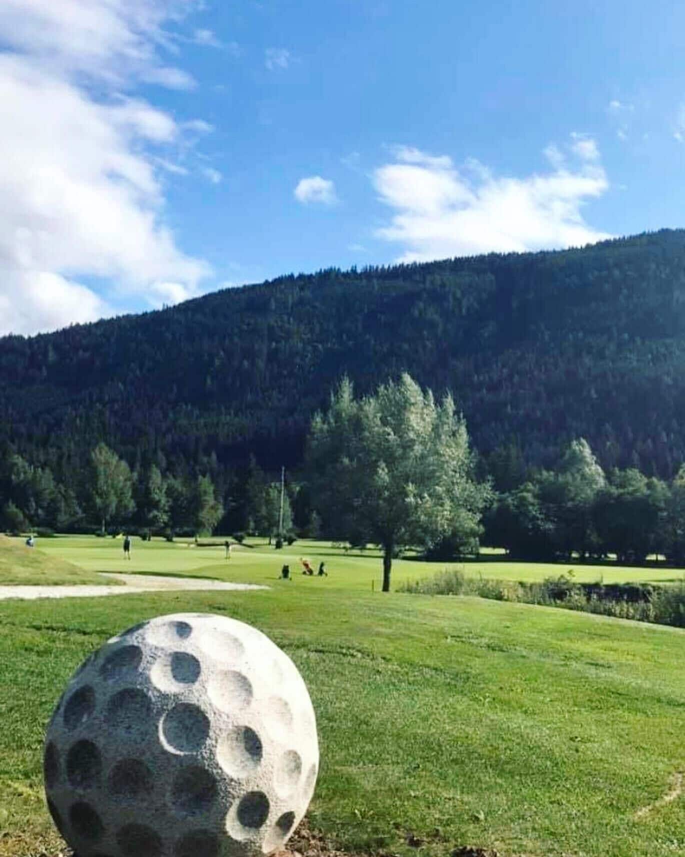 A golf course with a large golf ball in the foreground, a clear sky, and a forested hill in the background.