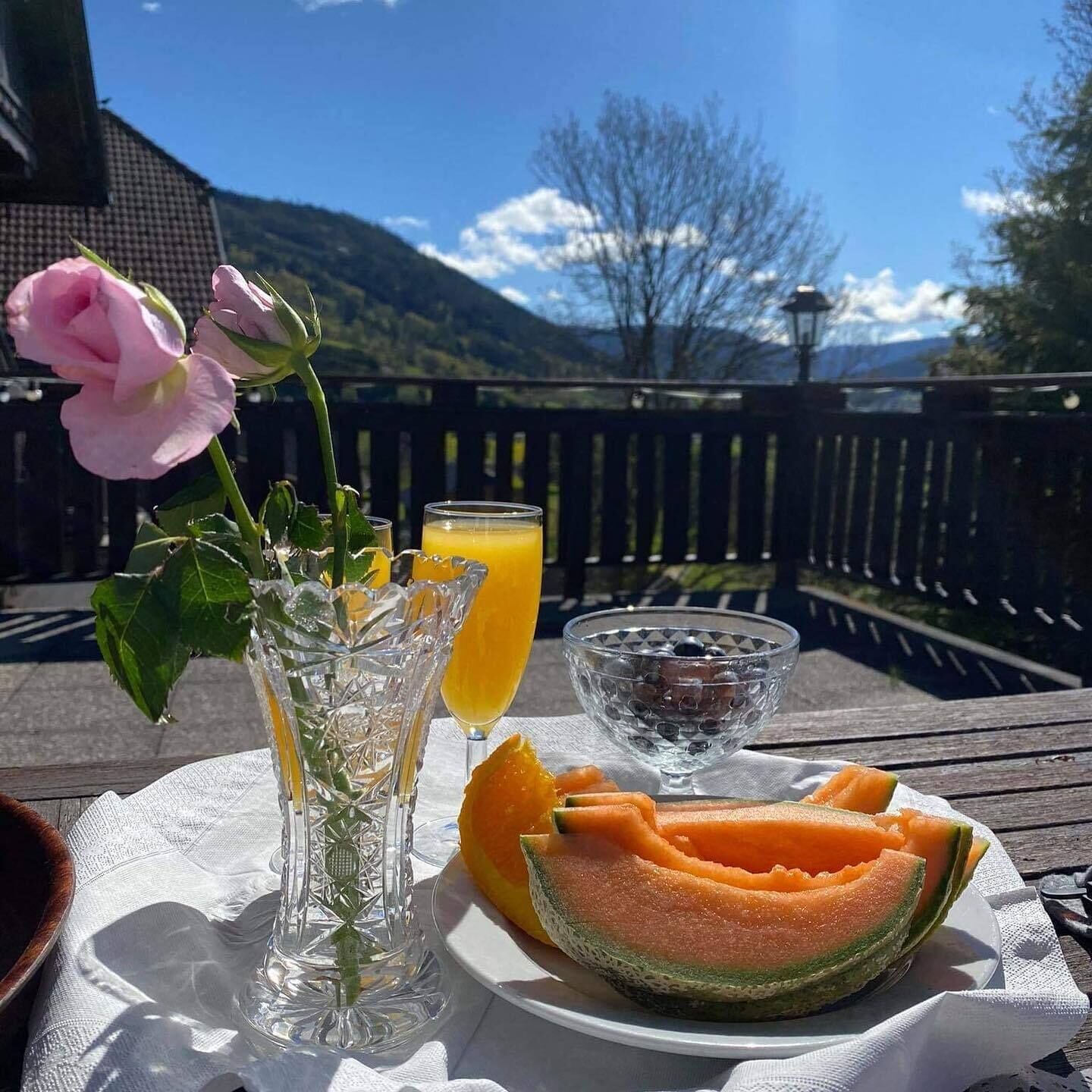 A table with a plate of cantaloupe, a glass of orange juice, and a glass of water on a wooden deck with a mountain view in the background.