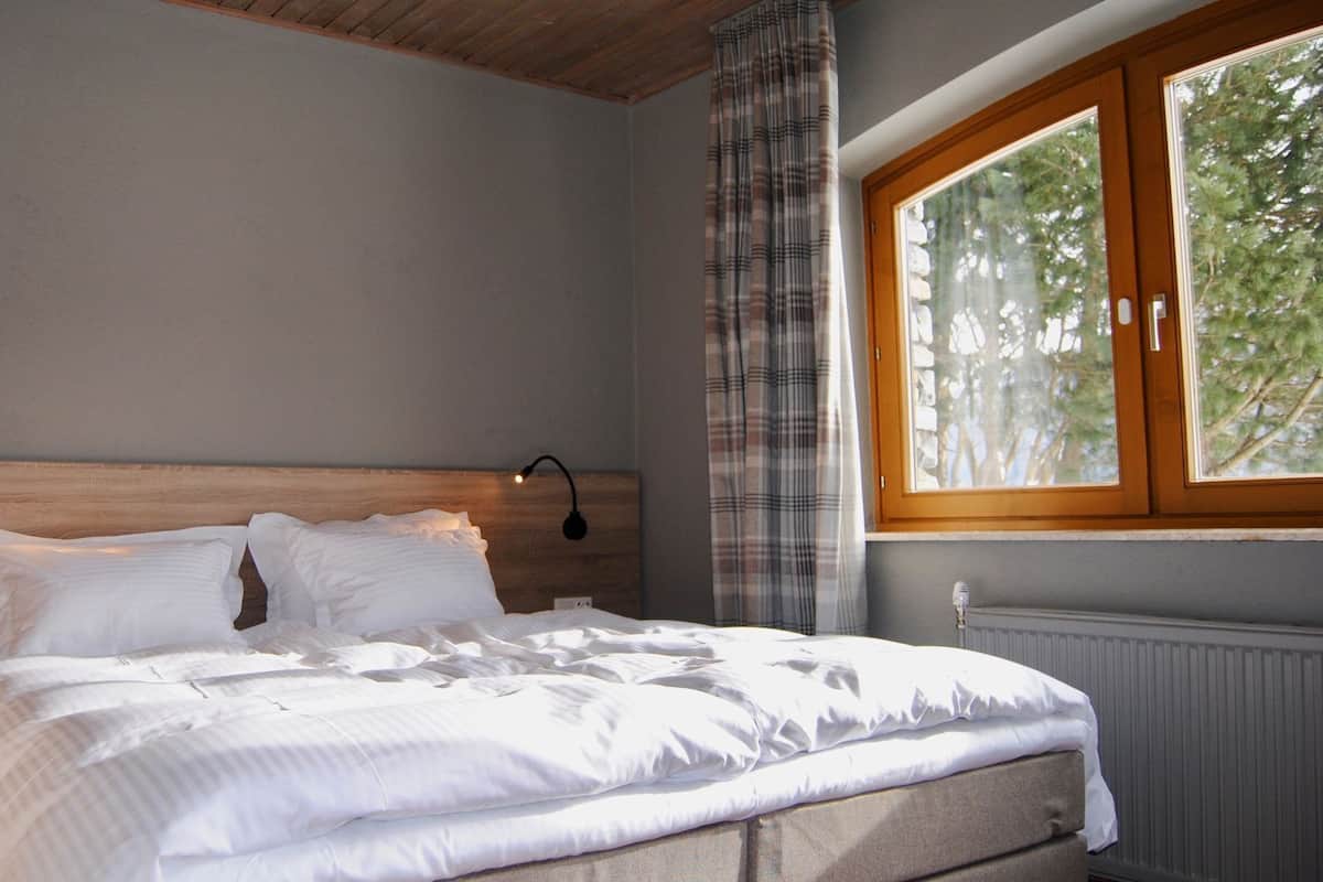 Una cama bien hecha con ropa blanca en una habitación con cabecera de madera, una ventana con vistas a árboles y una lámpara fijada a la pared.