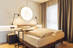 Habitación de hotel moderna con una cama grande, lámparas de noche, una lámpara de pared redonda y vistas al exterior a través de cortinas translúcidas.